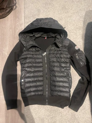 Monclear cardigan -  Snygg svart pufferjacka med huva och quiltad front. Jackan har dragkedja framtill, ribbade ärmar och sidor samt en liten ficka med logga på ärmen. Perfekt för kyliga dagar och enkel att styla till olika outfits.