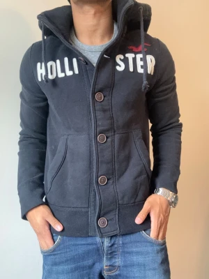 Vintage Hollister Cardigan Med Knappar - Väldigt sällsynt och vintage Hollister hoodie med knappar | Tröjan är i mycket bra skick| Längd ≈ 63cm, Bredd armhåla-amrhåla ≈ 53cm | Modell 175cm, 68kg | Hör av vid ytterligare frågor och priset kan diskuteras vid snabbt köp! |