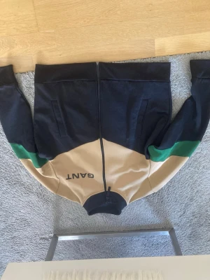 GANT ziptröja i svart, beige och grönt - Snygg ziptröja från GANT i svart, beige och grönt. Tröjan har hel dragkedja, hög krage och två fickor framtill. GANT-loggan syns på bröstet. Perfekt för en sportig och avslappnad stil.