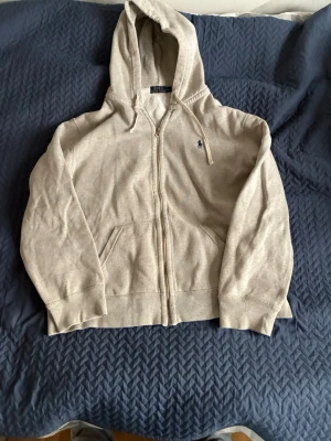 Beige hoodie från Polo Ralph Lauren - Snygg beige hoodie från Polo Ralph Lauren med dragkedja framtill och klassisk huva. Tröjan har två fickor på magen och den ikoniska lilla loggan broderad på bröstet. Perfekt för en avslappnad och stilren look.