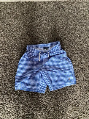 Blårutiga shorts med snörning - Snygga blå och vita rutiga shorts med snörning i midjan. Perfekta för sommaren och har en avslappnad passform. Klassisk design med små rutor och diskret logga framtill.