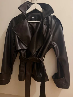 Svart trenchcoat i skinnimitation - Snygg svart trenchcoat från Bik Bok i skinnimitation med bred krage, knytskärp i midjan och dekorativa axelklaffar. Jackan har stora slag, knappar och uppvikta ärmslut för en trendig look. Perfekt för dig som gillar edgy och stilrena outfits.