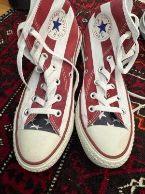 Converse All Star USA high tops - Snygga Converse All Star high tops med USA-flagga. Röd, vit och blå canvas med vita snören och gummisula. Klassisk rund tå och Converse-logga på sidan. Perfekta för dig som vill sticka ut med en unik sneaker.