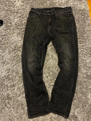 Svarta jeans från Replay - Säljer ett par svarta jeans från Replay med klassisk femficksmodell och raka ben. Jeansen har diskreta slitningar och en liten röd detalj på bakfickan. Tillverkade i mjukt denimtyg som sitter snyggt och bekvämt. Den är 93 cm lång och 36 cm bredd 