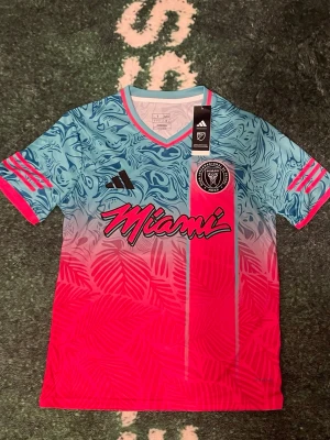 Inter Miami limited edition blå/rosa🌸🦋 - Snygg Inter Miami matchtröja från Adidas i blått och rosa med grafiskt mönster och klubbmärke på bröstet. Tröjan har korta ärmar, V-ringning och 'Miami' tryckt på framsidan. Perfekt för fotboll och still. Strl S. Extra pris om du tar 2 eller fler.