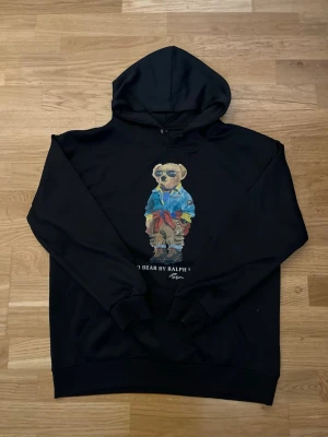 Svart hoodie Polo Bear Ralph Lauren - Svart hoodie med huva från Ralph Lauren med den ikoniska Polo Bear-trycket framtill. Björnen har solglasögon, blå jacka och färgglada detaljer. Hoodien har ribbade muddar och känguruficka. Perfekt för en avslappnad och trendig stil. Lappar och sånt är dock avklippta.