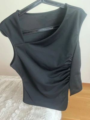 Svart asymmetrisk topp med rynk - Säljer en svart ärmlös topp med asymmetrisk skärning och rynkdetalj på ena sidan. Toppen har en unik form med snedskuren nederkant och bred axel. Materialet ser ut att vara stretchigt och mjukt, perfekt för en trendig look.