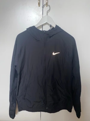 Svart vindjacka med huva från Nike - Snygg svart vindjacka från Nike med reflekterande logga på bröstet och detaljer på ärmarna. Jackan har dragkedja framtill, två sidofickor och en praktisk huva. Tillverkad i lätt och vindtätt material, perfekt för blåsiga dagar.