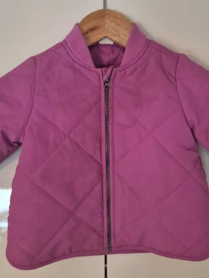 Lila quiltad jacka med dragkedja - Snygg lila quiltad jacka med dragkedja framtill och ribbad krage. Jackan har ett enkelt, stilrent mönster med rutiga sömmar och långa ärmar. Perfekt för kyligare dagar och lätt att matcha med olika outfits.i storlek 74 är 