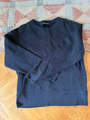 Svart sweatshirt från Acne Studios - En stilren svart sweatshirt från Acne Studios med rund halsringning och ribbade muddar vid ärmslut och nederkant. Tillverkad i mjukt bomullsmaterial som känns skönt mot huden. Perfekt för en clean och avslappnad look.