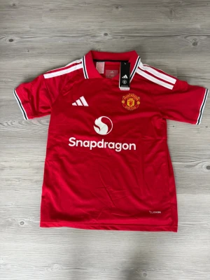 Manchester United matchtröja röd - Snygg röd Manchester United matchtröja från Adidas med klubbmärke och Snapdragon-logga på bröstet. Tröjan har vita detaljer på axlar och krage samt korta ärmar. Perfekt för fotbollsfans som vill visa sitt stöd på matchdagar.