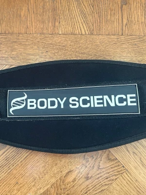 Svart Body Science träningsbälte - Säljer ett svart träningsbälte från Body Science, använd 1-2 ggr. Bältet har en bred, vadderad ryggdel och justerbart spänne i svart läder för extra stöd vid tunga lyft. Finns även kardborre för att snabbt ta på/av.
