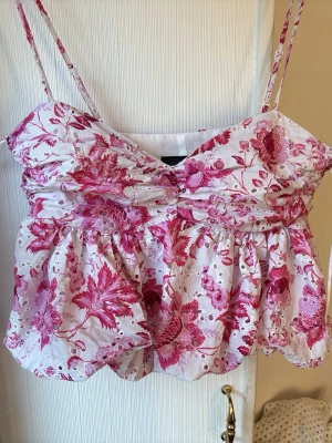 Blommig vit och rosa linnetopp - Supersöt linnetopp med tunna axelband och volangig nederdel. Toppen är vit med rosa blommigt mönster och har broderade detaljer som ger en romantisk känsla. Perfekt för varma dagar och somriga outfits. Aldrig använd!