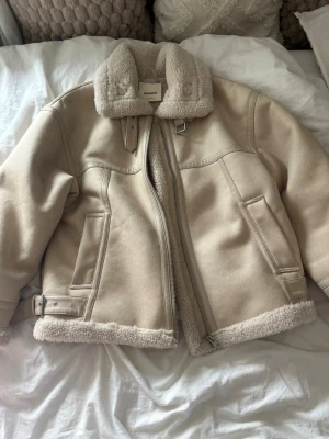 Höst och vinterjacka  - Jacka från PULL&BEAR. Beige jacka med vitt teddyfoder inuti. Spännen på båda sidorna och vid kragen. I stolek M passar bra till höst och vintern