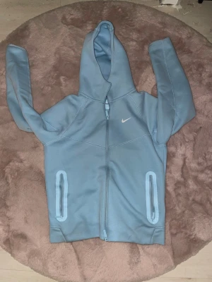 Ljusblå Nike hoodie med dragkedja - Snygg ljusblå hoodie från Nike med dragkedja framtill och två fickor med ljusblå detaljer. Klassisk huva och diskret Nike-logga på bröstet. Perfekt för en avslappnad och sportig stil.