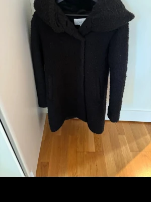 Svart ullkappa med stor krage - Snygg svart kappa i ullmix med stor, mysig krage och rak passform. Kappan har diskreta fickor på sidorna och stängs framtill. Perfekt för kyliga dagar och ger en stilren look.
