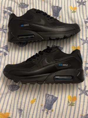 Svarta Nike Air Max sneakers - Säljer ett par svarta Nike Air Max sneakers med blå detaljer och synlig luftbubbla i sulan. Skorna har en klassisk låg siluett, robust yttersula och är tillverkade i en mix av läder och syntetmaterial. Perfekta för dig som gillar streetwear och sportig stil.