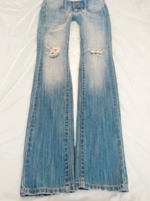 Ljusblå bootcut jeans med slitningar - Säljer ett par ljusblå bootcut jeans med slitningar och hål på låren. Jeansen har klassisk femficksmodell, låg midja och slitna detaljer både fram och bak. Tillverkade i denim med snygg tvättad look och vida ben. Midjemått:40 innerbenslängd: 84. Säljer pågrund av att dem inte  passar. 