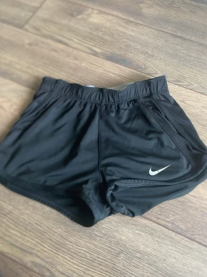Svarta träningsshorts från Nike - Snygga svarta träningsshorts från Nike med klassisk vit logga på ena benet. Tillverkade i lätt och snabbtorkande Dri-FIT-material som andas. Shortsen har elastisk midja och en sportig passform som passar perfekt till träning eller löpning.