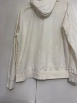 Vit hoodie med dragsko - En stilren vit hoodie med huva och dragsko. Hoodien har långa ärmar, bred mudd vid ärmslut och nederkant samt en avslappnad passform. Perfekt för en chill och enkel look. Materialet ser ut att vara mjukt och bekvämt, troligen bomull eller bomullsblandning.