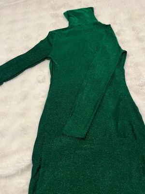 Grön glittrig långklänning polo - Snygg långklänning i glittrande grön färg med polokrage och långa ärmar. Klänningen har en figurnära passform och är perfekt för dig som vill sticka ut. Materialet känns mjukt och stretchigt, vilket gör den bekväm att bära.