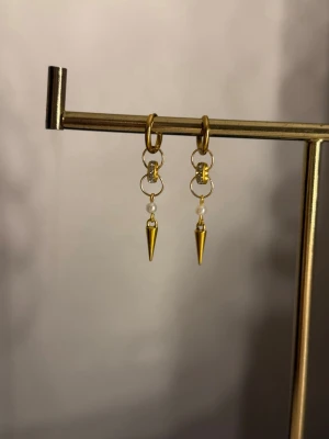 Guldiga örhängen med pärldetalj och spets - Säljer ett par trendiga örhängen i guldfärg med små vita pärlor och spetsiga hängen. Örhängena har en cool kedjedesign och kombinerar metall med pärldetaljer för en edgy look. Perfekta för dig som gillar statement-accessoarer.