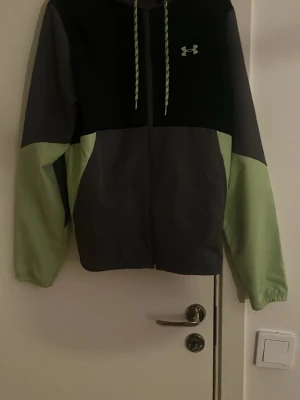 Grå och grön hoodie från Under Armour - Snygg hoodie från Under Armour i grått och ljusgrönt med dragkedja framtill. Hoodien har en huva med snören och logga på bröstet. Tillverkad i ett mjukt och bekvämt material, perfekt för chill eller träning.