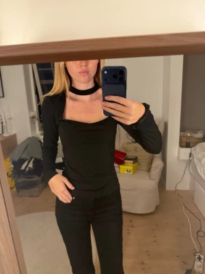 Svart långärmad topp med choker - Trendig svart långärmad topp med fyrkantig urringning och fastsydd choker-detalj runt halsen. Toppen har en tight passform och är perfekt för dig som vill ha en stilren men edgy look. Materialet känns mjukt och stretchigt.