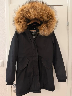 Jacka - Snygg svart parkas med stor luva kantad av brun päls. Jackan har dragkedja och knappar framtill, två stora fickor med lock och ribbade muddar vid ärmsluten. Sparsamt använd. Det går att ta bort pälsen. Pris går att diskutera💕