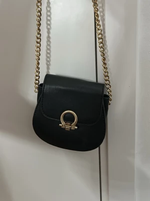 Svart axelväska med guldkedja - Snygg svart axelväska i rundad form med guldfärgad kedjerem och dekorativt guldfärgat spänne framtill. Väskan är tillverkad i slätt material och har en stilren, trendig look som passar till många outfits.