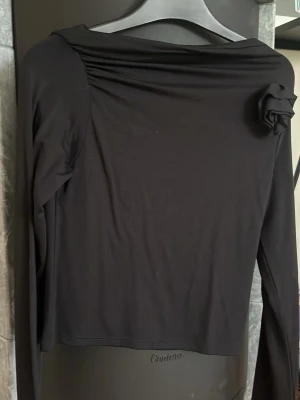 Svart långärmad topp med rosettdetalj - Stilren svart långärmad topp med en snygg rosettdetalj på axeln. Toppen har en något vid halsringning och är figurnära i modellen.
