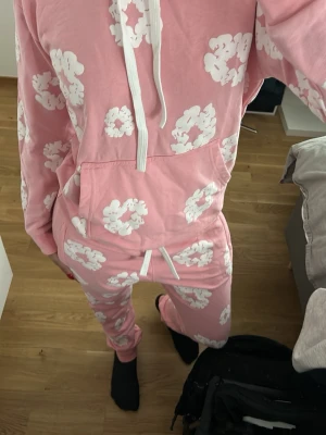Rosa loungewear set med blommor - Mysigt loungewear set i rosa med vita blommönster. Setet består av en hoodie med dragsko och magficka samt matchande byxor med resår och snörning i midjan. Perfekt för chill dagar hemma eller när du vill vara extra bekväm.
