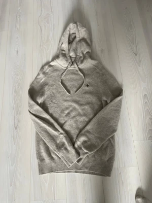Beige hoodie från Morris - Snygg beige hoodie från Morris i mjukt stickat material. Klassisk modell med huva och dragsnören samt diskret Morris-logga på bröstet. Perfekt för dig som gillar stilrena och bekväma plagg med en exklusiv touch.