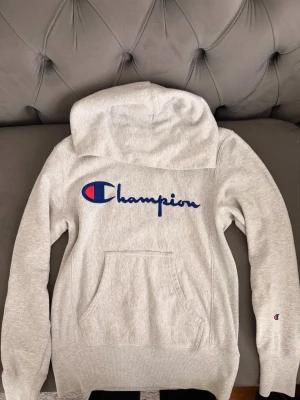Grå hoodie från Champion - Snygg grå hoodie från Champion med stor logga broderad i blått och rött på bröstet. Klassisk känguruficka framtill och liten logga på ärmen. Mjuk bomullsblandning och avslappnad passform med stor huva.