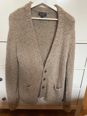 Beige stickad kofta Polo Jeans  - Beige stickad kofta från Polo Jeans Company, tillverkad av italienskt garn. Koftan har djup V-ringning, ribbstickade muddar, knäppning framtill med tre knappar och två små fickor. Perfekt för lager-på-lager och chill stil.