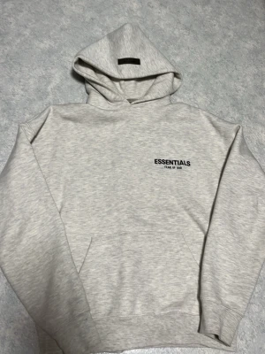 Ljusgrå Essentials hoodie XXS - Säljer en ljusgrå hoodie från Essentials Fear of God i storlek XXS. Hoodien har huva, magficka och svart tryck på bröstet. Tillverkad i mjukt och bekvämt material, perfekt för en avslappnad streetwear-look.