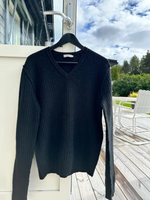 Svart stickad tröja från J.Lindeberg - Klassisk svart stickad tröja från J.Lindeberg med V-ringning och ribbad struktur. Tröjan har långa ärmar och en enkel, stilren design som passar perfekt till många olika outfits. Materialet ser mjukt och bekvämt ut.