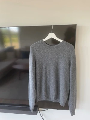 Grå stickad tröja med rund hals från Uniqlo - En enkel och stilren grå stickad tröja med rund hals och långa ärmar. Tröjan har ribbade muddar vid ärmslut och nederkant, perfekt för lager på lager under kyliga dagar. Passar till det mesta och är ett måste i garderoben. Tveka inte på att höra av dig vid frågor!