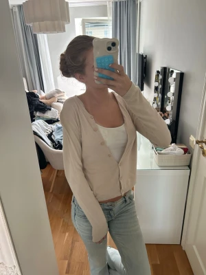 Beige basic kofta med knappar - En enkel och stilren beige kofta med v-ringning och knäppning framtill. Koftan har långa ärmar och är gjord i ett tunt, mjukt material som passar perfekt till jeans eller kjol. Snygg att bära både öppen och stängd.