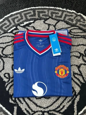 Blå Manchester United tröja Adidas - Snygg blå Manchester United matchtröja från Adidas med röda och vita detaljer. Tröjan har klubbmärke, Adidas-logga och Snapdragon-tryck på bröstet. Klassisk V-ringad krage och tre röda ränder på axlarna. Perfekt för fotboll eller streetwear.