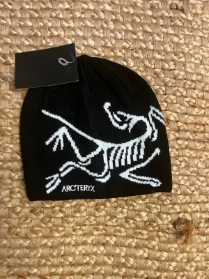 Black Arc’teryx Beanie - Svart stickad mössa från Arc'teryx med stor vit logga i form av ett skelett på framsidan. Klassisk passform och mjukt material som sitter skönt på huvudet. Perfekt för kalla dagar och outdoor-aktiviteter.