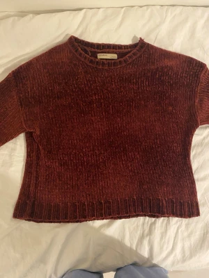 Roströd chenilletröja med rund hals - Mysig stickad tröja i roströd chenille med rund halsringning och ribbade muddar vid ärmslut och nederkant. Tröjan har en boxig passform och långa ärmar, perfekt för kyliga dagar. Materialet ger en mjuk och glansig känsla.