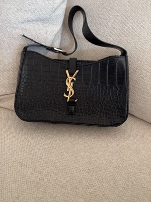 Svart handväska från YSL - Snygg svart handväska i croco-präglat läder från YSL med guldfärgad logga framtill. Väskan har axelrem och stilren form som ger en lyxig känsla. Perfekt accessoar för dig som gillar exklusiv design och trendiga detaljer.