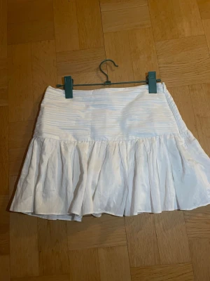 Vit volangkjol med dragkedja - Snygg vit kortkjol med volangkant och rynkade detaljer upptill. Kjolen har en diskret dragkedja i sidan och är tillverkad i ett lätt, glansigt material som ger en fräsch look. Perfekt för varma dagar och somriga outfits.