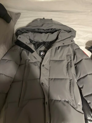 Grå dunjacka från Canada Goose - Säljer en grå dunjacka från Canada Goose med stor huva och klassisk logga på ärmen. Jackan har rejäla fickor framtill, dragkedja och knappar samt en puffig, varm design perfekt för kalla dagar. Materialet är slitstarkt och jackan är riktigt isolerande.