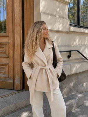 Beige kappa med knytskärp från NA-KD - Stilren beige kappa från NA-KD i storlek 36. Kappan har bred krage, knytskärp i midjan och mjukt, ull-liknande material. Perfekt för lager på lager och ger en clean look till alla outfits.