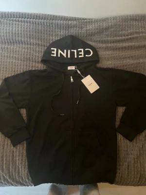 Svart hoodie med dragkedja från CELINE - Svart hoodie från CELINE med vit logga på huvan. Hoodien har dragkedja framtill, två stora fickor och justerbar dragsko i huvan. Tillverkad i mjukt material och har ribbade muddar vid ärmslut och nederkant. Perfekt för en avslappnad och trendig look.