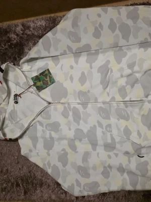 Vit Bape  hoodie med dragkedja - Säljer en vit hoodie med grått och ljusgult camomönster, hel dragkedja och huva. På huvan finns broderade detaljer med hajmotiv och bokstäverna 'WGM' i vitt och svart. Hoodien har en streetwear-vibe och är perfekt för dig som gillar unika plagg.