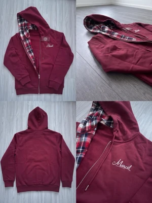 Vinröd hoodie med rutigt foder - Snygg vinröd hoodie med dragkedja och huva. Insidan av huvan har ett rutigt foder i rött, vitt och svart. På bröstet finns en broderad text 'Marcell'. Hoodien har ribbade muddar och fickor framtill.