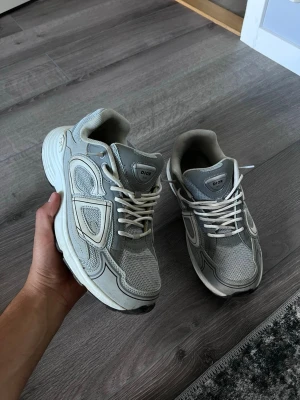 Dior b30 - Snygga gråa Dior sneakers med en chunky sula och detaljer i både mesh och läder. Skorna har vita snören och en sportig vibe med tydliga paneler och logga på plösen. Perfekta för dig som gillar streetwear och vill sticka ut med exklusiva detaljer.